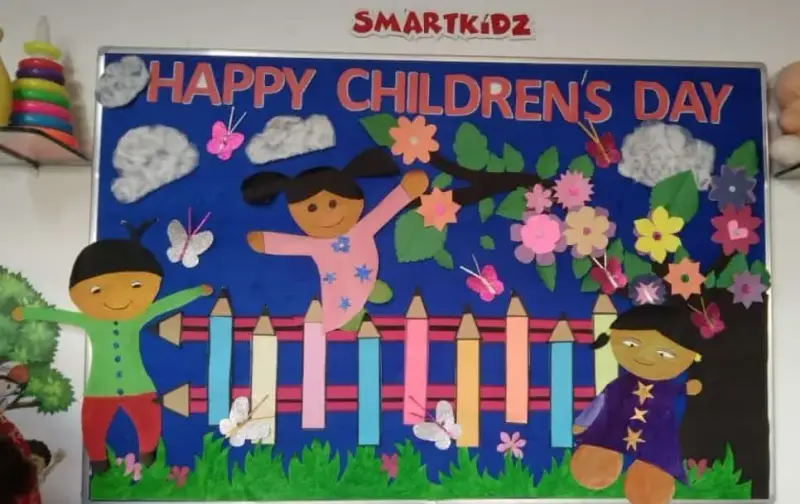A classroom display board reading Happy Children's Day, with paper children, flowers, butterflies, and a pencil fence.
